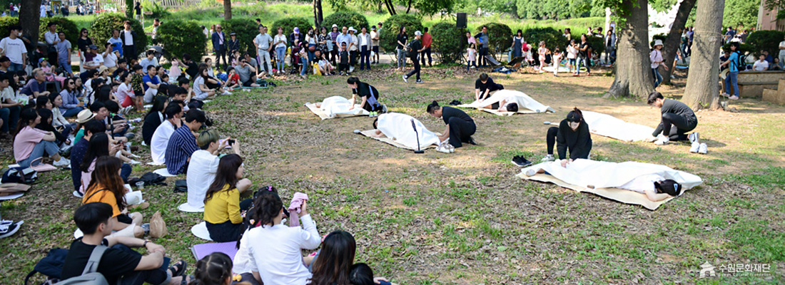 공원에서 관객들이 둘러앉은 가운데, 흰 천에 덮여 누워있는 사람들을 검은 옷의 공연자들이 돌보는 퍼포먼스. (선택된 큰 사진)