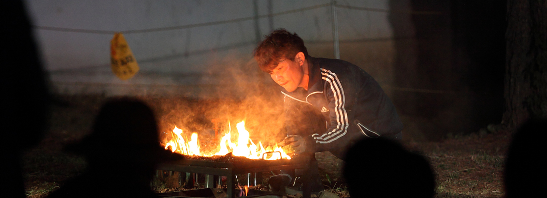 한 남성이 밤에 활활 타오르는 화로 위로 몸을 숙여 불을 들여다보고 있다. (선택된 큰 사진)