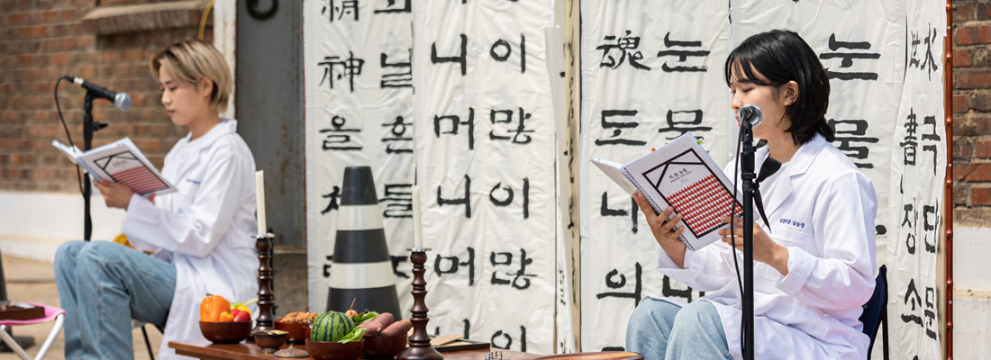 한글 서예가 적힌 현수막 앞에서 흰 가운을 입은 두 공연자가 책을 보며 낭독 퍼포먼스를 하고 있다. (선택된 큰 사진)