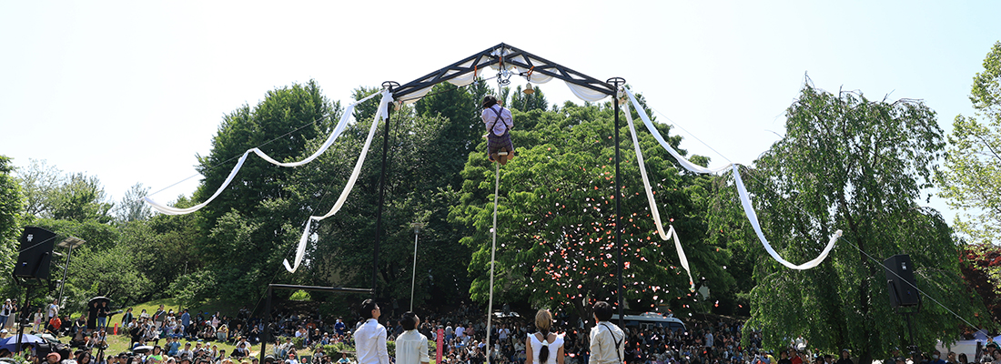 푸른 하늘을 배경으로 한 남성이 커다란 흰 천이 달린 장대를 들고 줄타기 공연을 하고 있다.