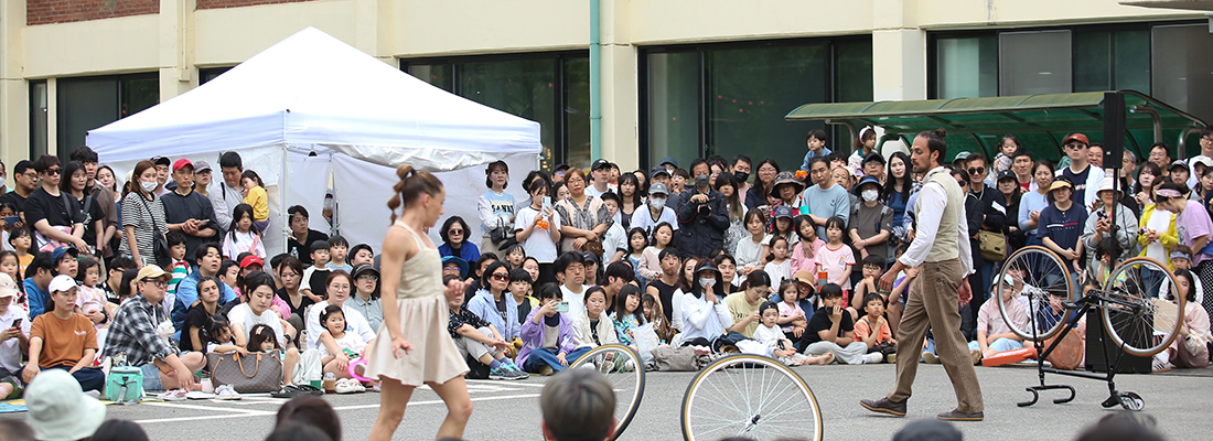 거리에서 조끼를 입은 공연자가 관객 한 명을 대상으로 재미있는 표정을 지으며 공연을 하고 있고, 많은 시민들이 이를 둘러싸고 구경하고 있다.